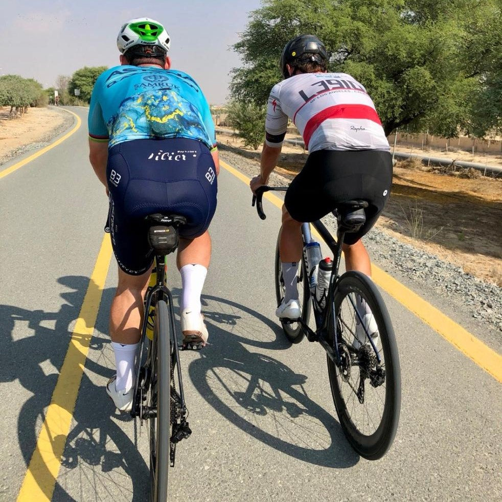 Two cyclists on the road wearing white cycling socks with a blue toe area, the socks reach mid-calf height.